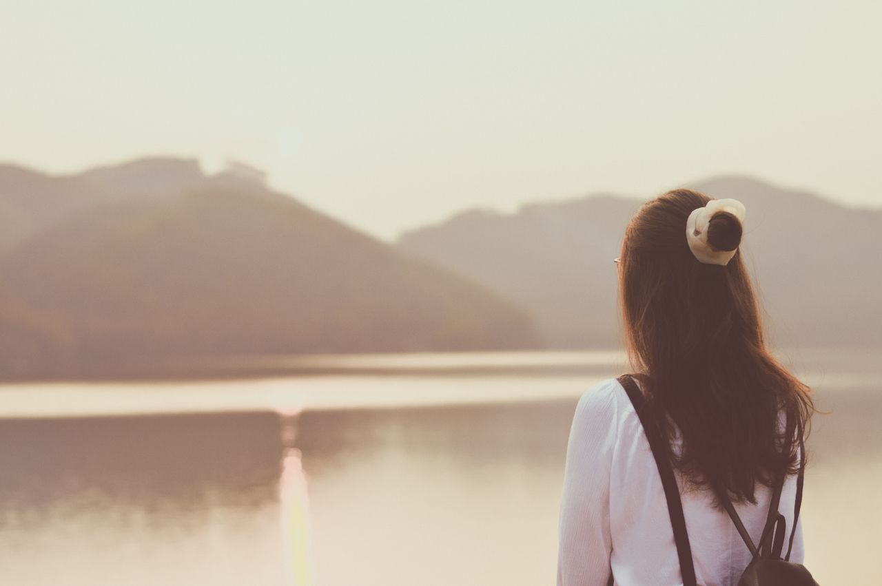 A woman gazes across a serene lake at sunset, surrounded by calm waters and distant hills.